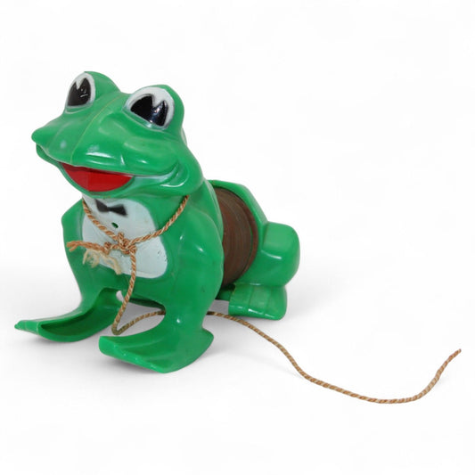 Vintage green plastic frog with pull-along rope and built-in cassette/spool tape mechanism. Frog wears a painted tuxedo bib and has a red smiling mouth. Pull the cord to wind the internal tape wheel and release it to watch the frog's "belly" spool slowly unwind, likely playing a jingling or recorded sound. Charming retro mechanical toy — a whimsical novelty from days gone by!
