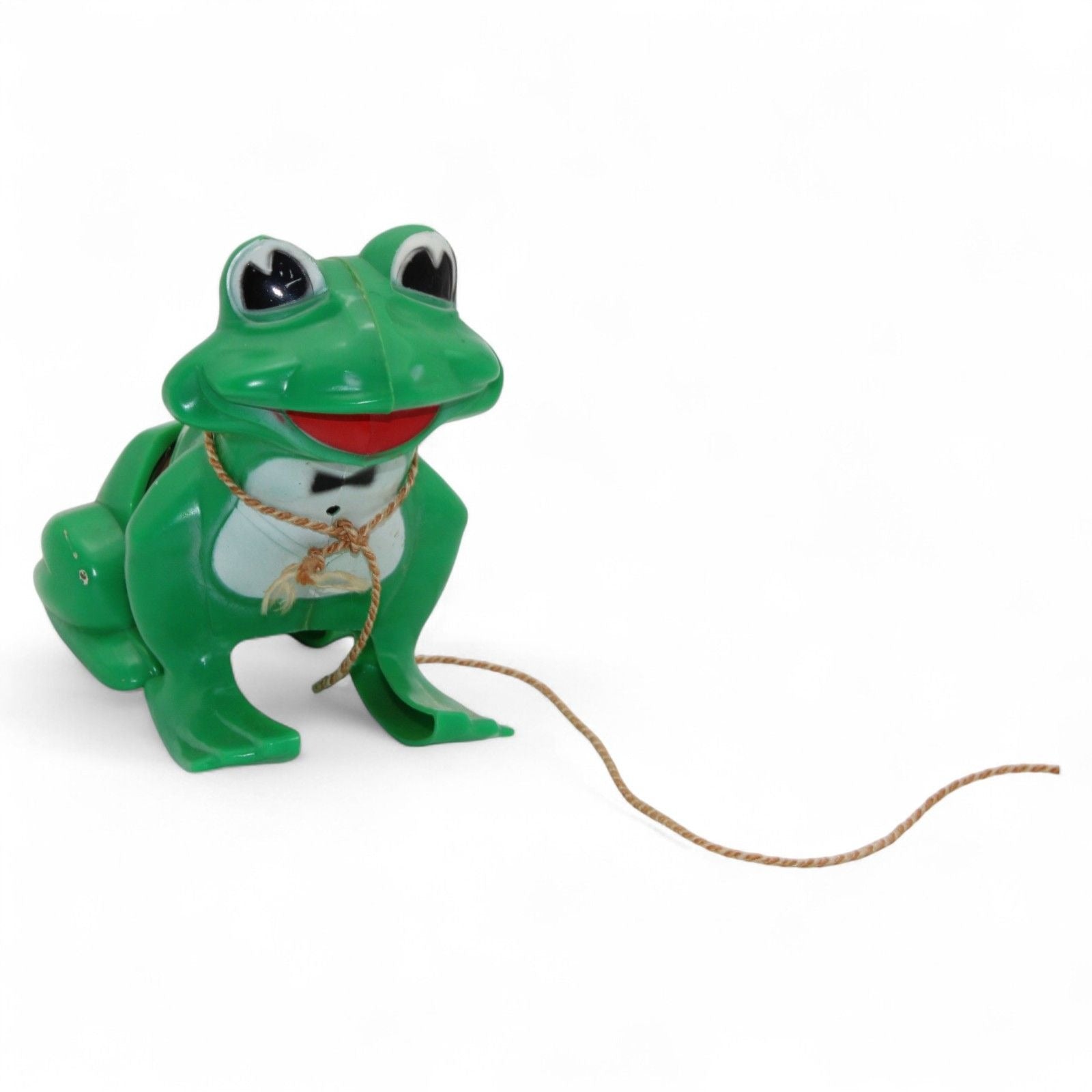 Vintage green plastic frog with pull-along rope and built-in cassette/spool tape mechanism. Frog wears a painted tuxedo bib and has a red smiling mouth. Pull the cord to wind the internal tape wheel and release it to watch the frog's "belly" spool slowly unwind, likely playing a jingling or recorded sound. Charming retro mechanical toy — a whimsical novelty from days gone by!
