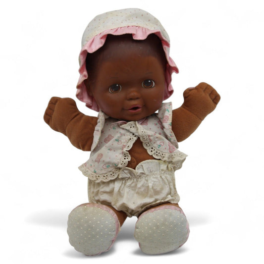 A vintage plush baby doll with a brown vinyl head, pink bonnet and lace-trimmed vest, featuring soft fabric arms and cloth bloomers.
